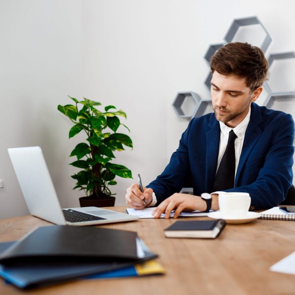 Young successful businessman in suit sitting at workplace, writing, office background.